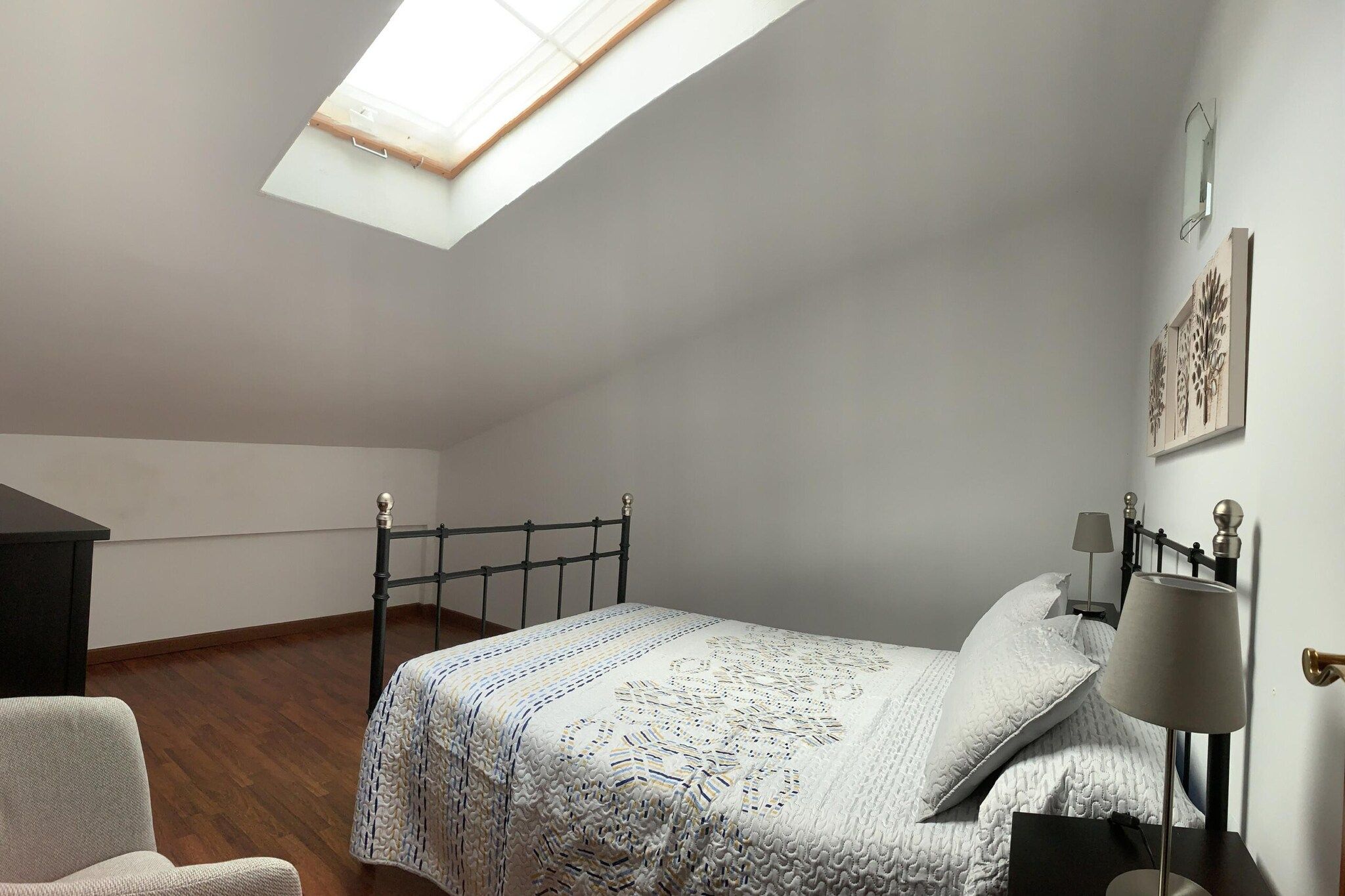 A bedroom with a skylight, a bed with a patterned bedspread, a bedside lamp, and a chair.
