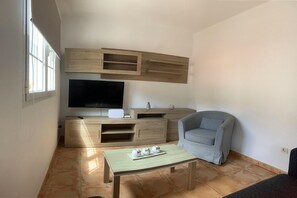 Een knus woongedeelte met een flatscreen televisie, houten meubels, een grijze fauteuil en een tafeltje met decoratieve objecten.