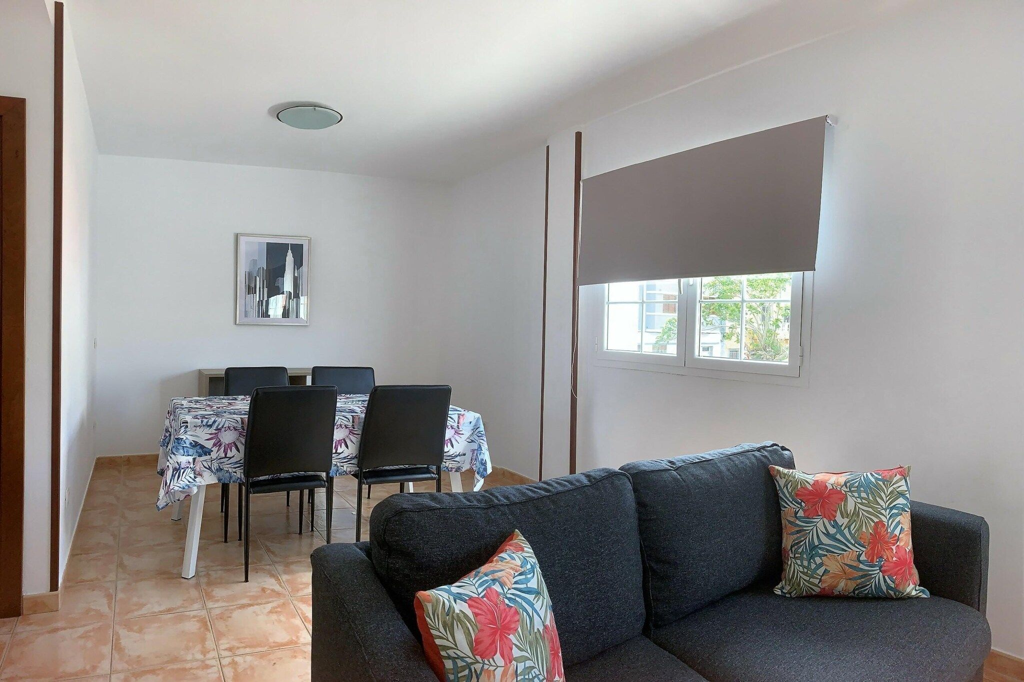 A living room with a sofa, two pillows, a dining table with chairs, and a cityscape painting on the wall.