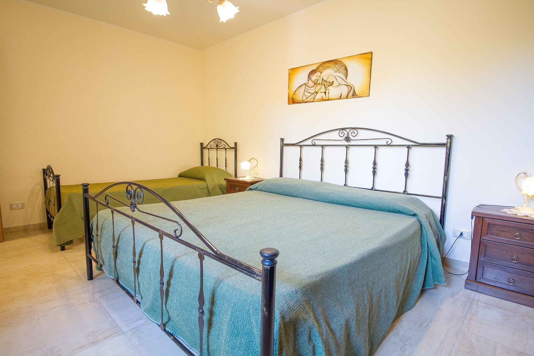 A bedroom with a metal bed frame, two pillows, a bedside table with a lamp, and a wall-mounted picture.