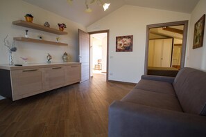 A living room with a sofa, wooden flooring, a kitchen area with cabinets and shelves, and a doorway leading to another room. - Beautiful Sea-View Attic Suite (Trecastagn)