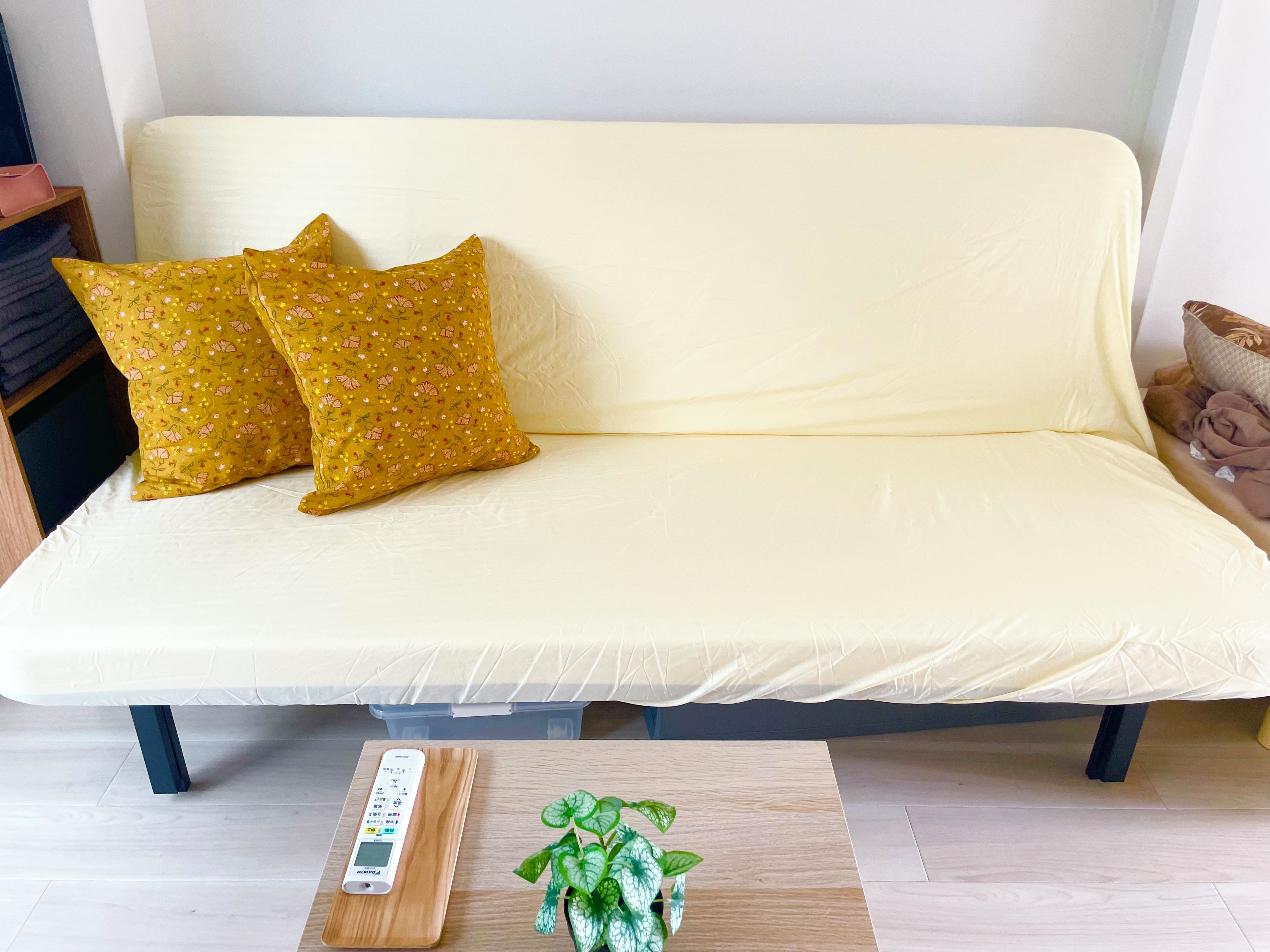 Ein Sofa mit gelber Hülle und zwei gemusterten Kissen, ein kleiner Holztisch mit Fernbedienung und einem Topf mit Pflanze, sowie eine weiße Wand im Hintergrund.
