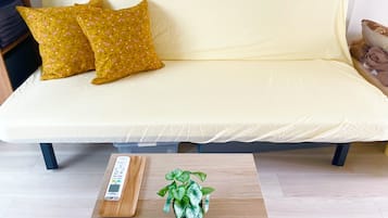 Ein Sofa mit gelber Hülle und zwei gemusterten Kissen, ein kleiner Holztisch mit Fernbedienung und einem Topf mit Pflanze, sowie eine weiße Wand im Hintergrund.