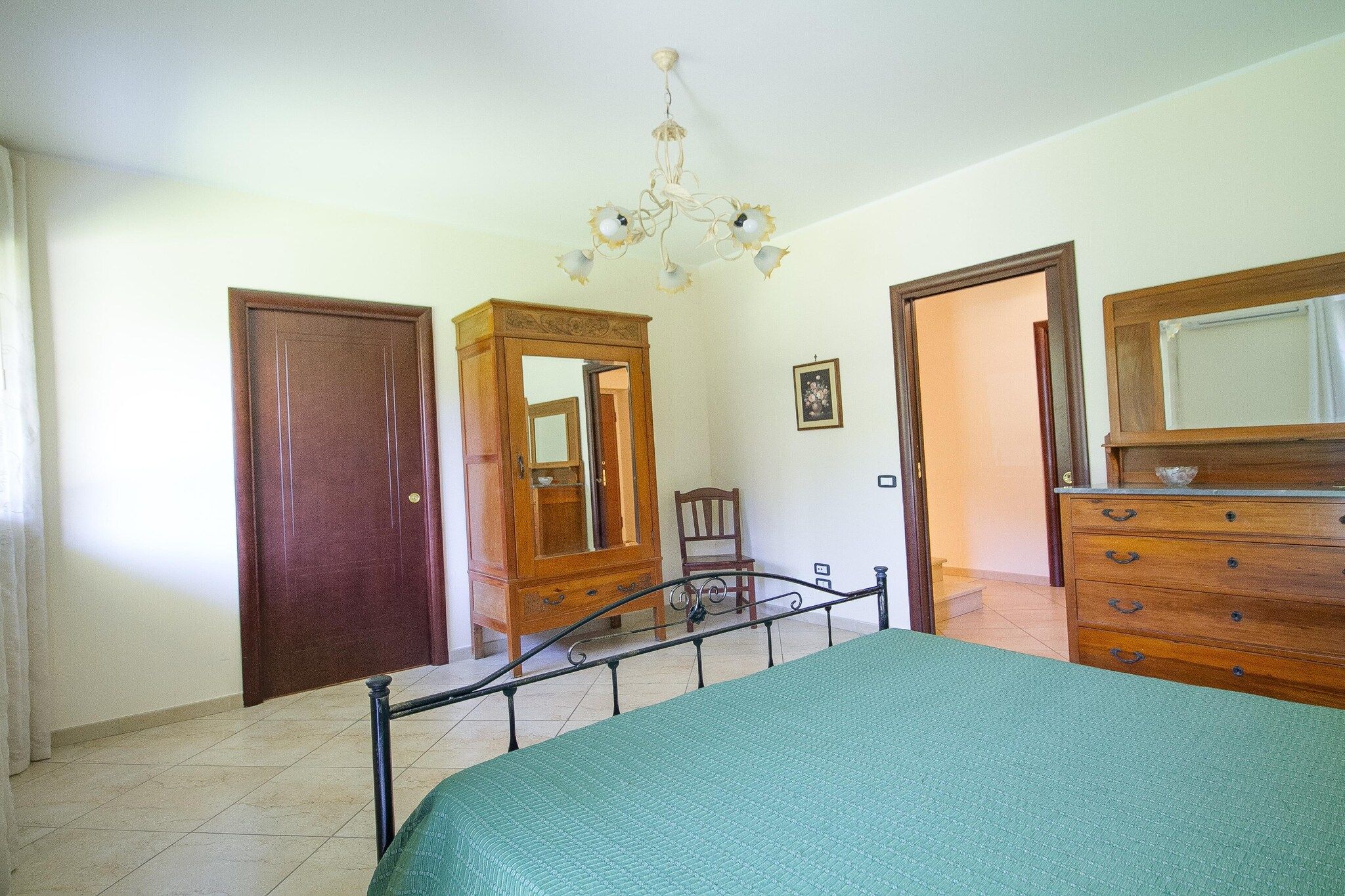 A bedroom with a bed, a dresser, a chair, a mirror, and a chandelier.