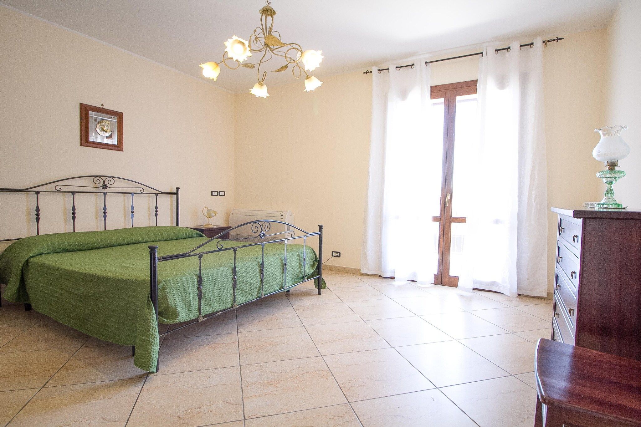 Een slaapkamer met een bed, een kroonluchter, een raam met gordijnen en een dressoir.