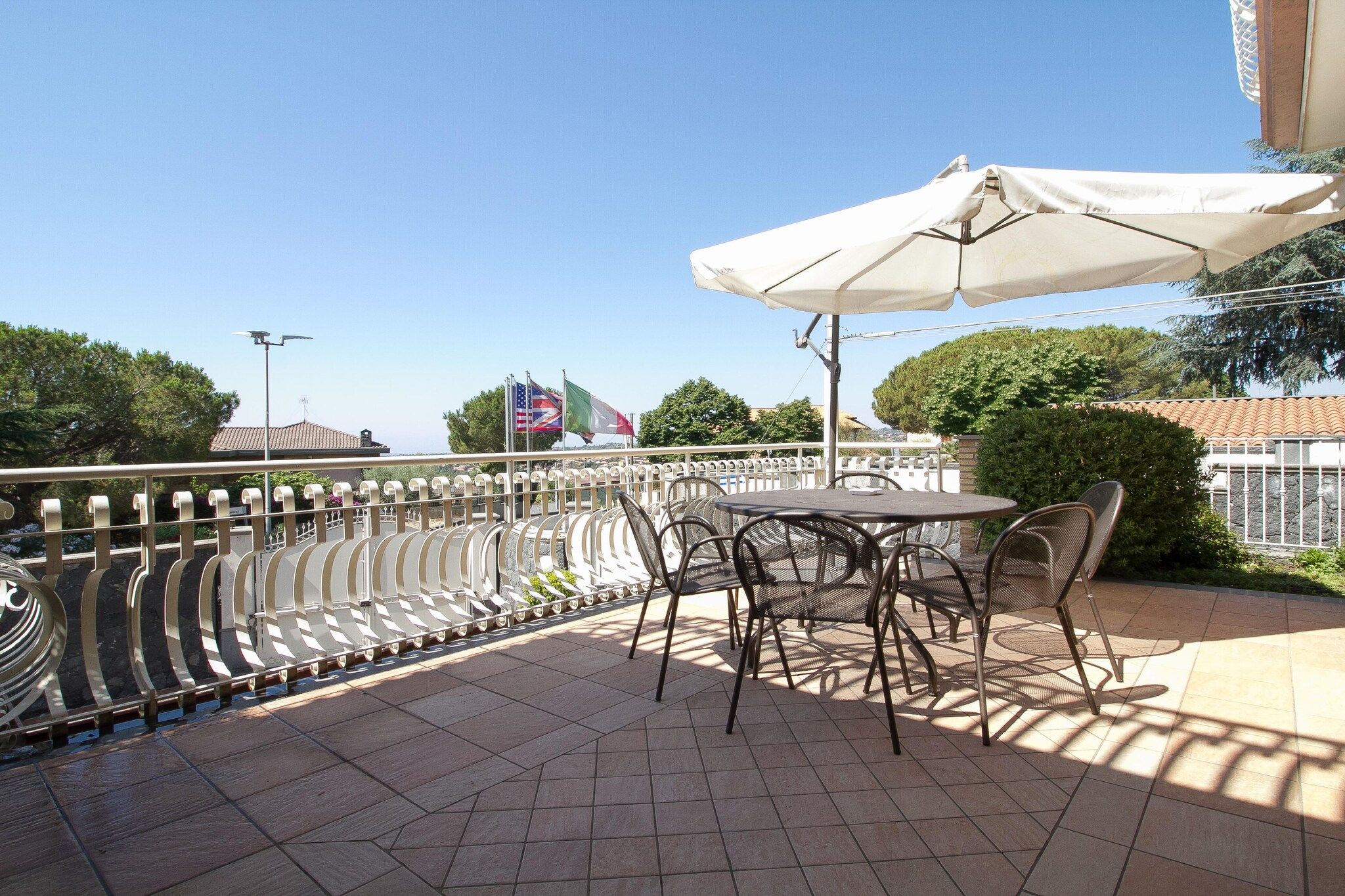 Eine Terrasse im Freien mit Tisch und Stühlen, einem weißen Sonnenschirm und Flaggen im Hintergrund.