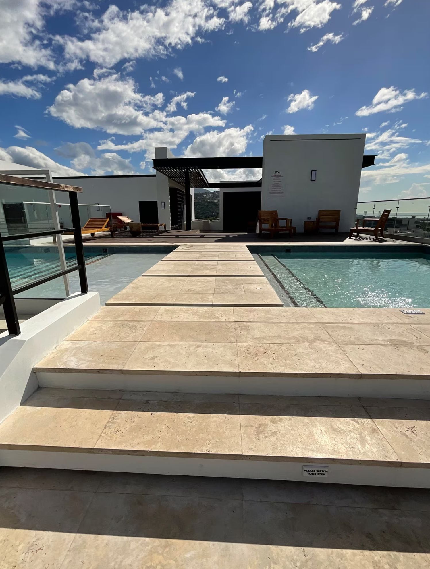Zona de piscina con escalones que conducen a un edificio, cielo despejado y vista al mar.