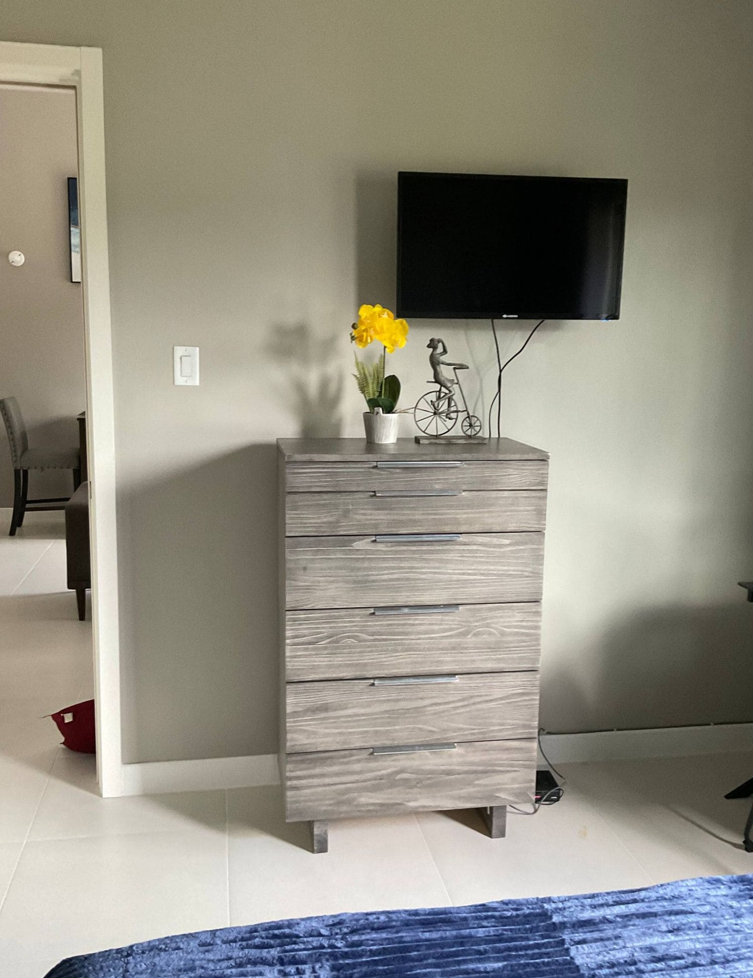 A room with a grey dresser, a flat-screen TV mounted on the wall, and a potted plant with yellow flowers on top.