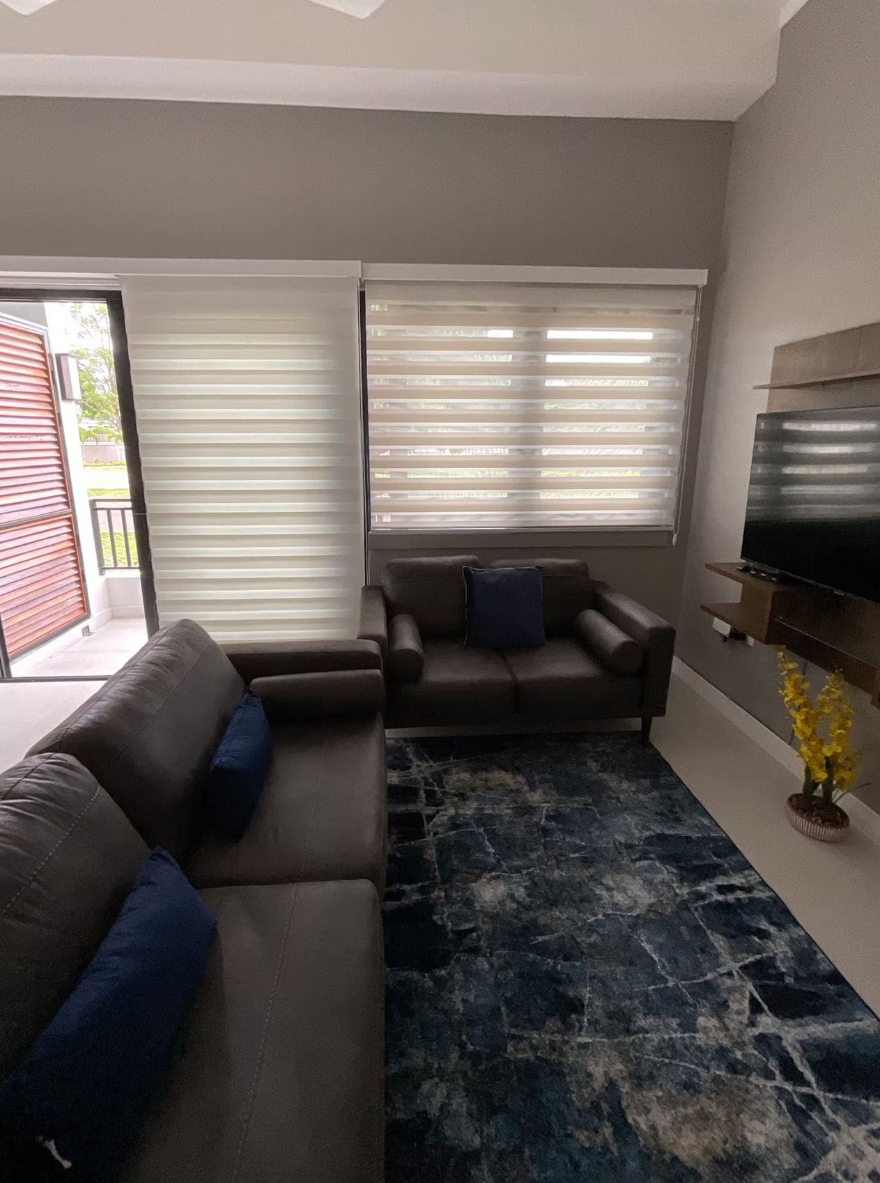 Een woonkamer met een hoekbank, een kleinere bank, een flatscreen televisie en een vloerkleed met een steenmotief.