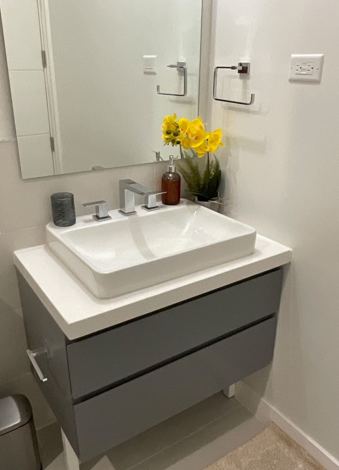 Un bany amb un lavabo blanc, un mirall, un suport per a tovalloles i una planta amb flors grogues.