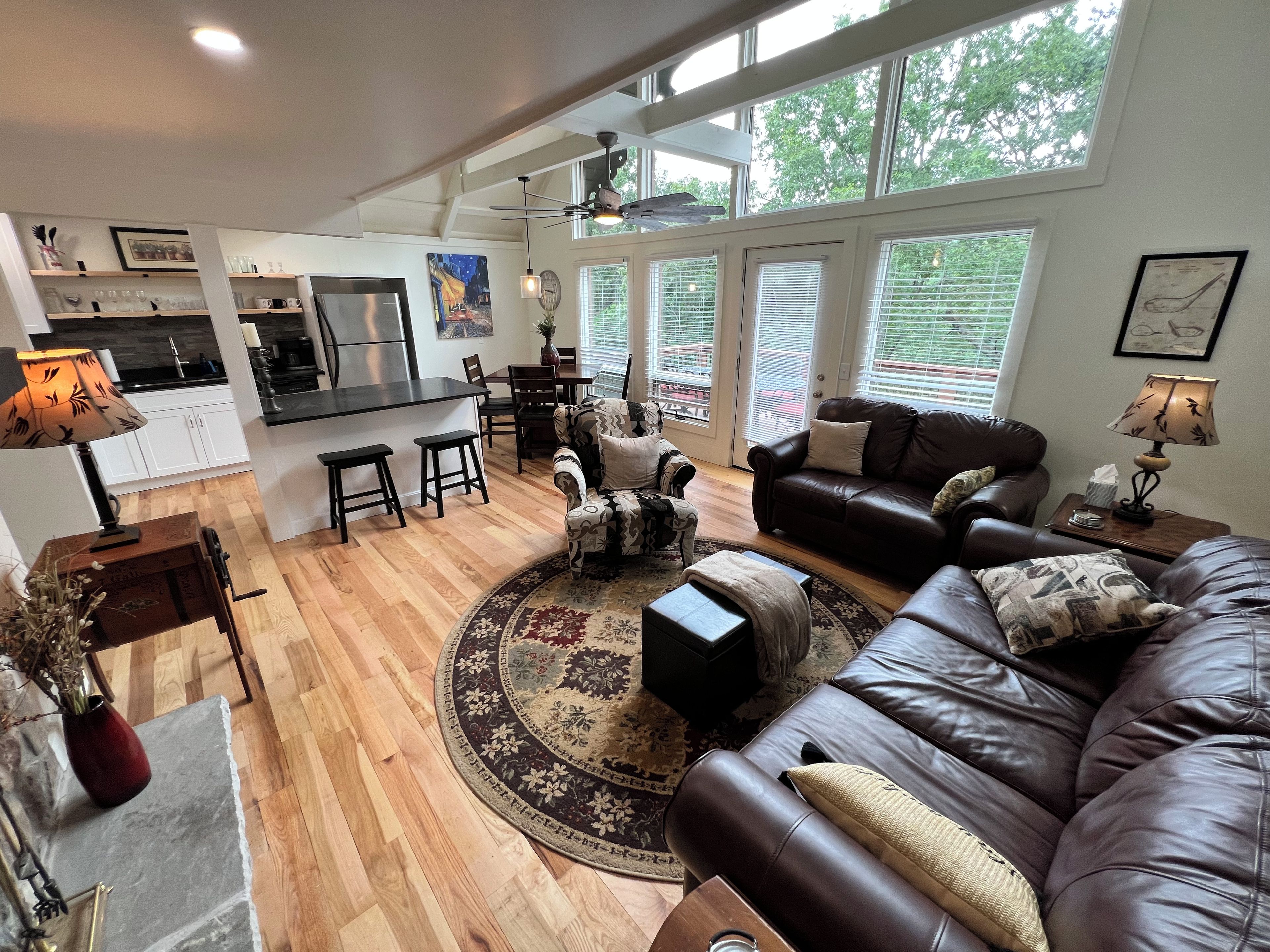 A spacious living area with a leather sofa, a coffee table, and a dining table with chairs. There is a kitchen area with a refrigerator, stove, and bar stools. The room has large windows with blinds and a ceiling fan.