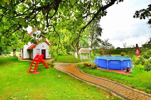 Un jardin résidentiel avec une structure d’escalade rouge, une maison blanche, une piscine bleue et un chemin pavé.