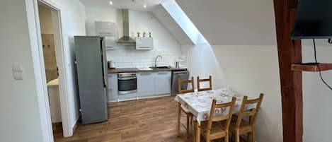 A compact kitchen with modern appliances, a dining area with wooden chairs, and a sloped ceiling with skylight.