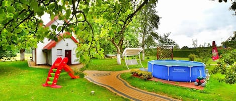 A residential garden with a white house, a red climbing frame, a blue swimming pool, and a paved pathway.