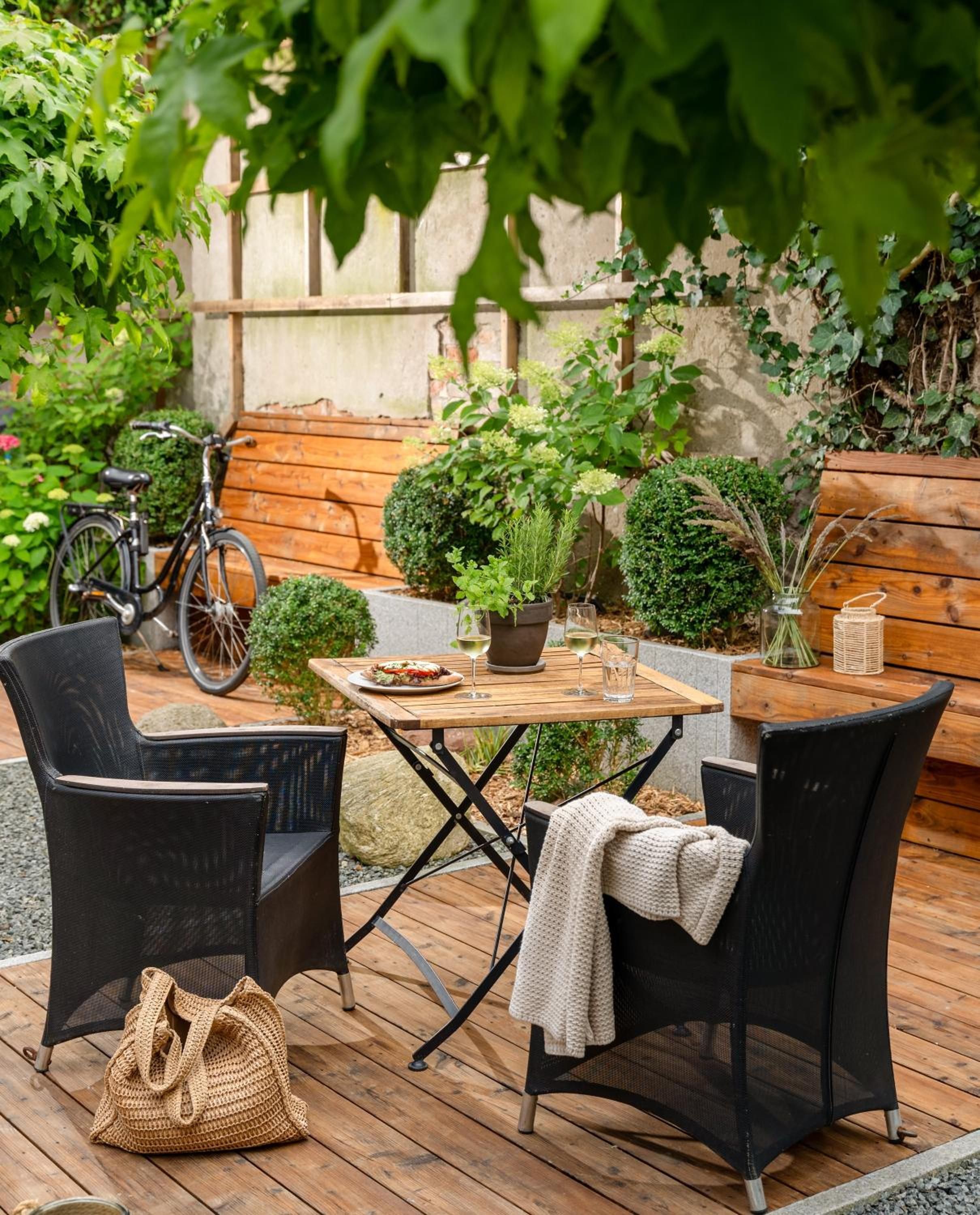 Une terrasse extérieure avec une table en bois, deux chaises, une plante en pot et un vélo en arrière-plan.