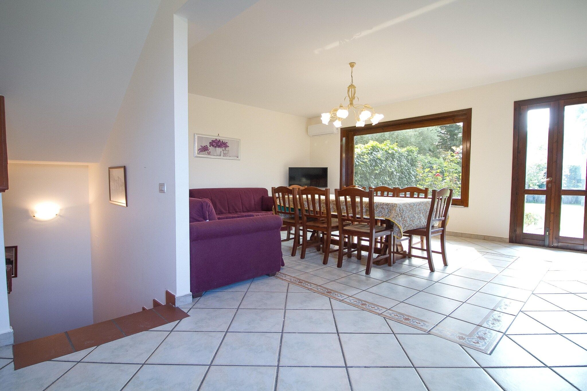 A living room with a purple sofa, wooden dining table, chairs, a TV, and a large window with a view of greenery.