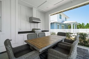 An outdoor patio with a wooden table and wicker chairs, a grill, and a view of a house. - 🌴Dune's Perch: Beach Club Resort with Pool + Hot Tub + On-Site Dining (Gulf Shores)