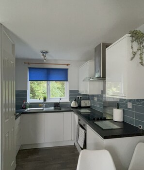A modern kitchen with white cabinets, a black countertop, and a window with blue curtains. - Cosy, Comfortable 3bd holiday cottage set on Tolroy, Hayle. (Tolroy Manor Hayle)