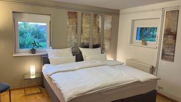 Ein Schlafzimmer mit einem großen Bett, einem Nachttisch, einem Sessel und einem Fenster mit Blick auf Grünflächen.