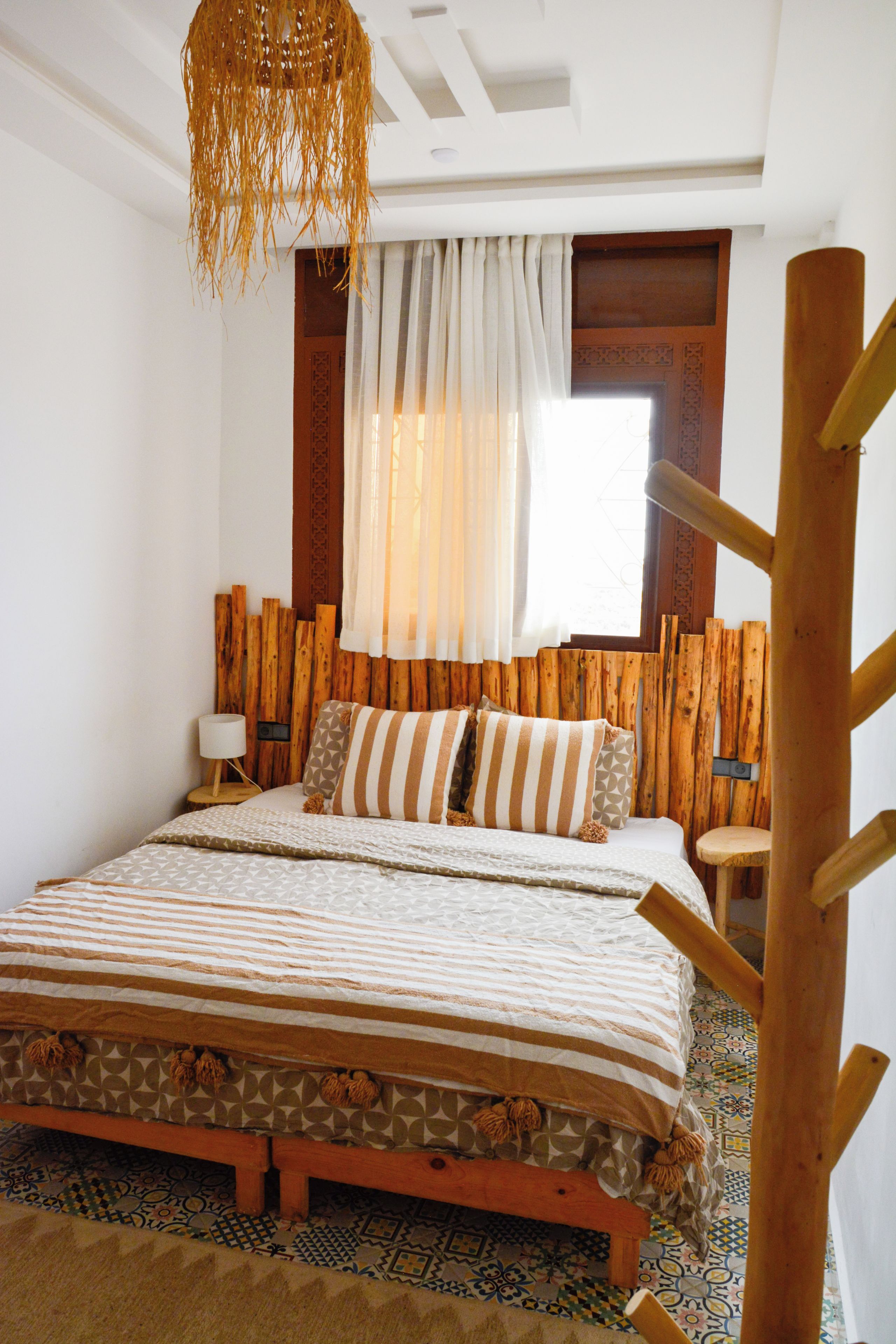 Un dormitorio con cabecera de madera, una cama con ropa de cama estampada, una mesita de noche con lámpara y una ventana con cortinas.
