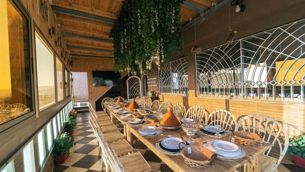 Une longue table à manger entourée de chaises, un sol à carreaux et des plantes suspendues.