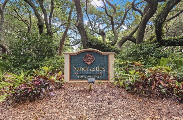 A sign for Sandcastles Villas 200-254, surrounded by lush greenery and trees.