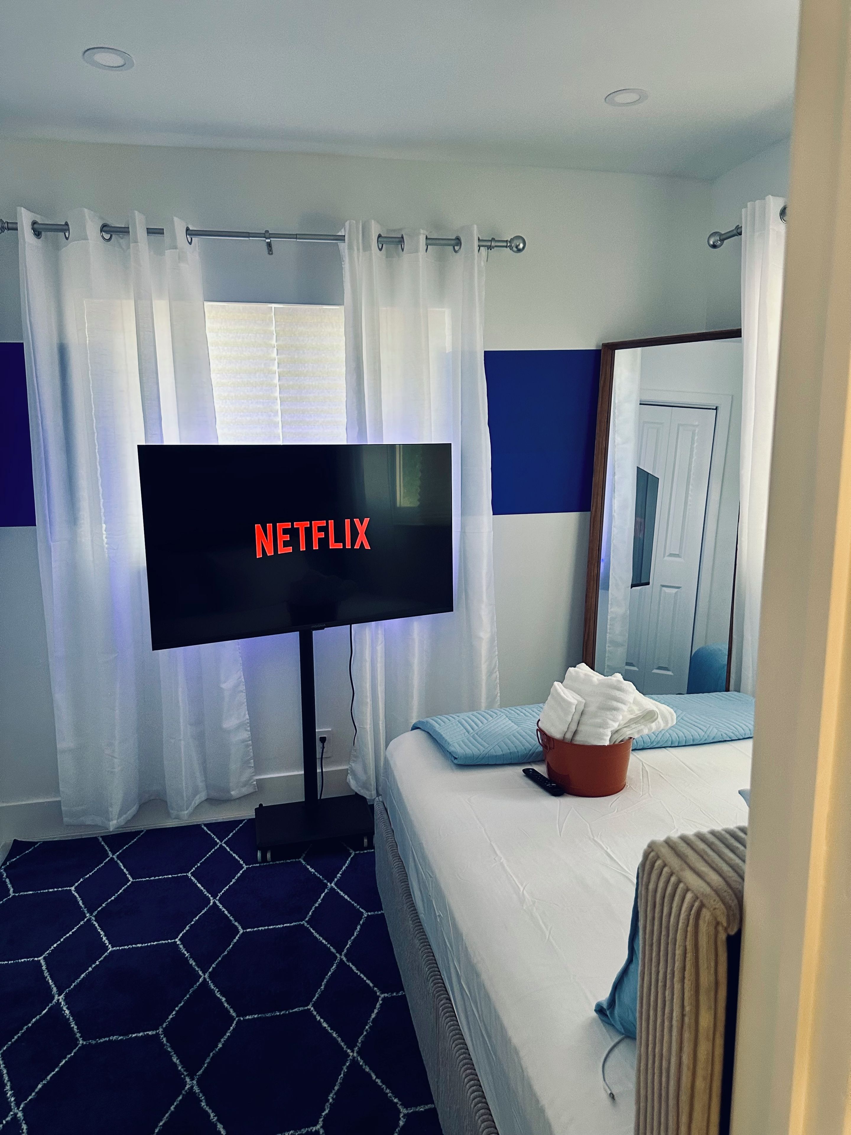 Ein Hotelzimmer mit einem Bett, einem Fernseher, der 'NETFLIX' anzeigt, einem Fenster mit Vorhängen und einem blauen Teppich mit sechseckigem Muster.