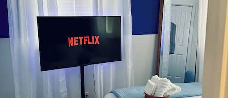 Ein Hotelzimmer mit einem Bett, einem Fernseher, der 'NETFLIX' anzeigt, einem Fenster mit Vorhängen und einem blauen Teppich mit sechseckigem Muster.