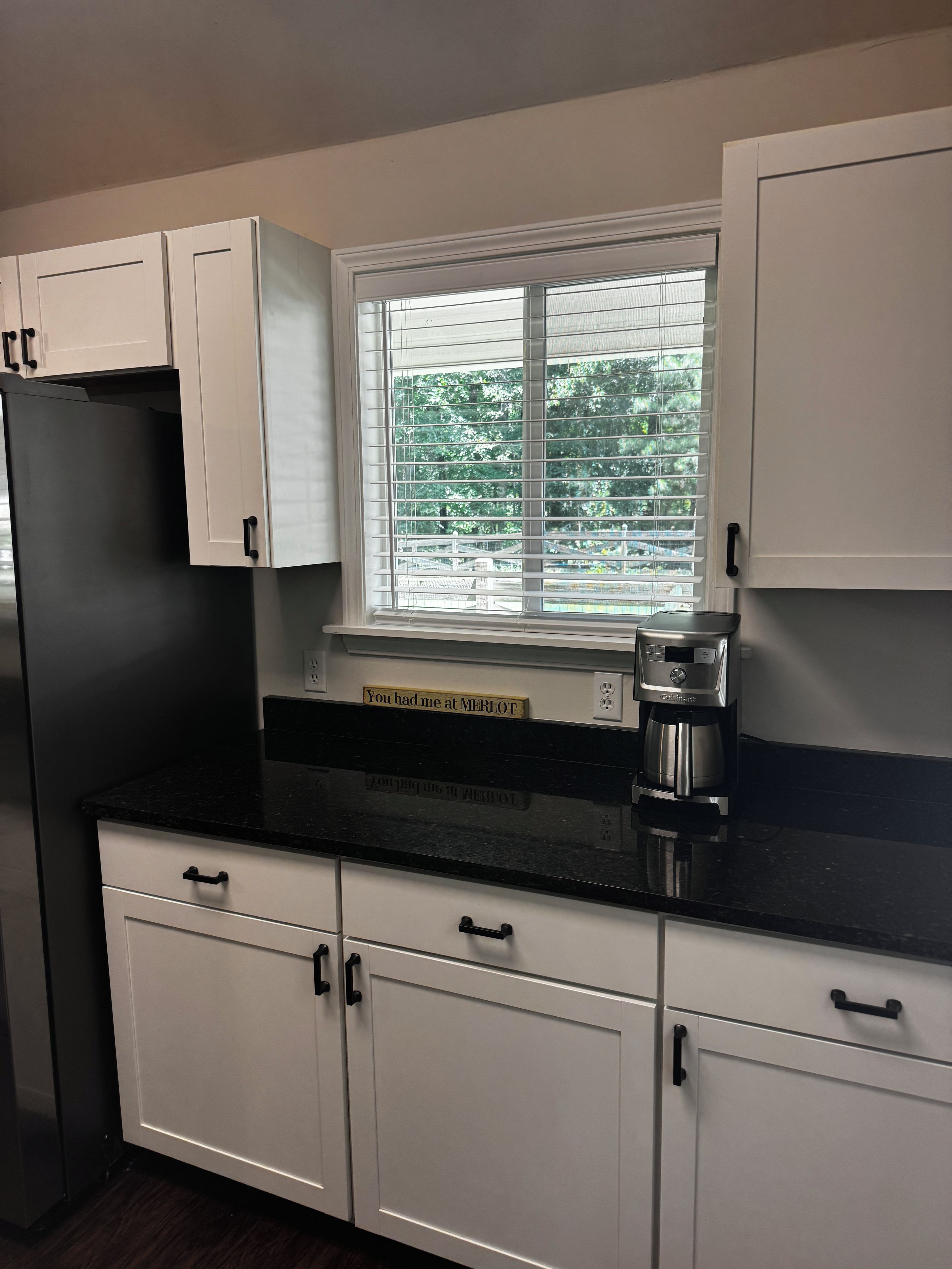 A kitchen with white cabinets, a black countertop, a window with blinds, and a coffee maker.
