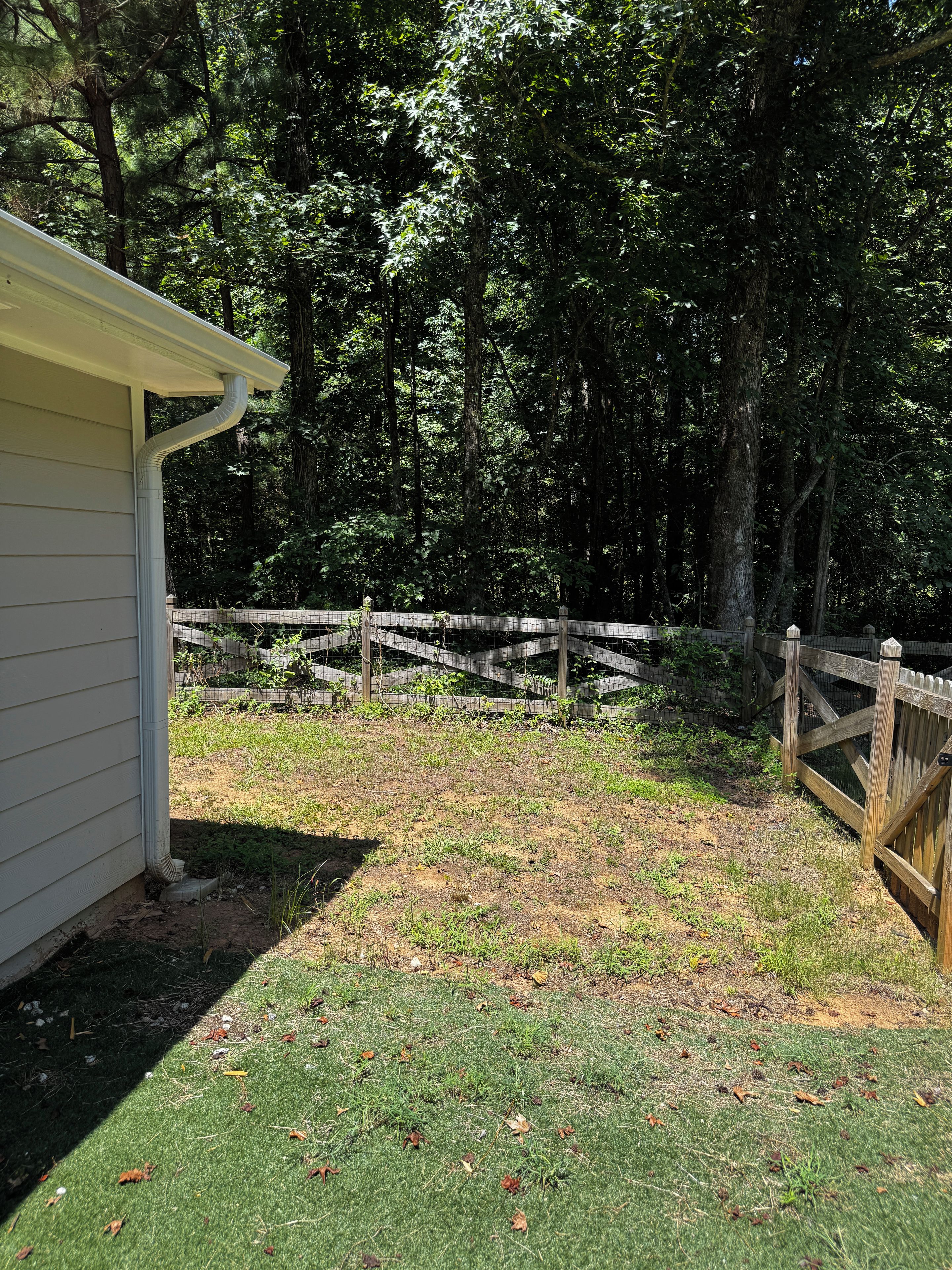 A fenced yard with a wooden gate, a grassy area, and a building with a white exterior.