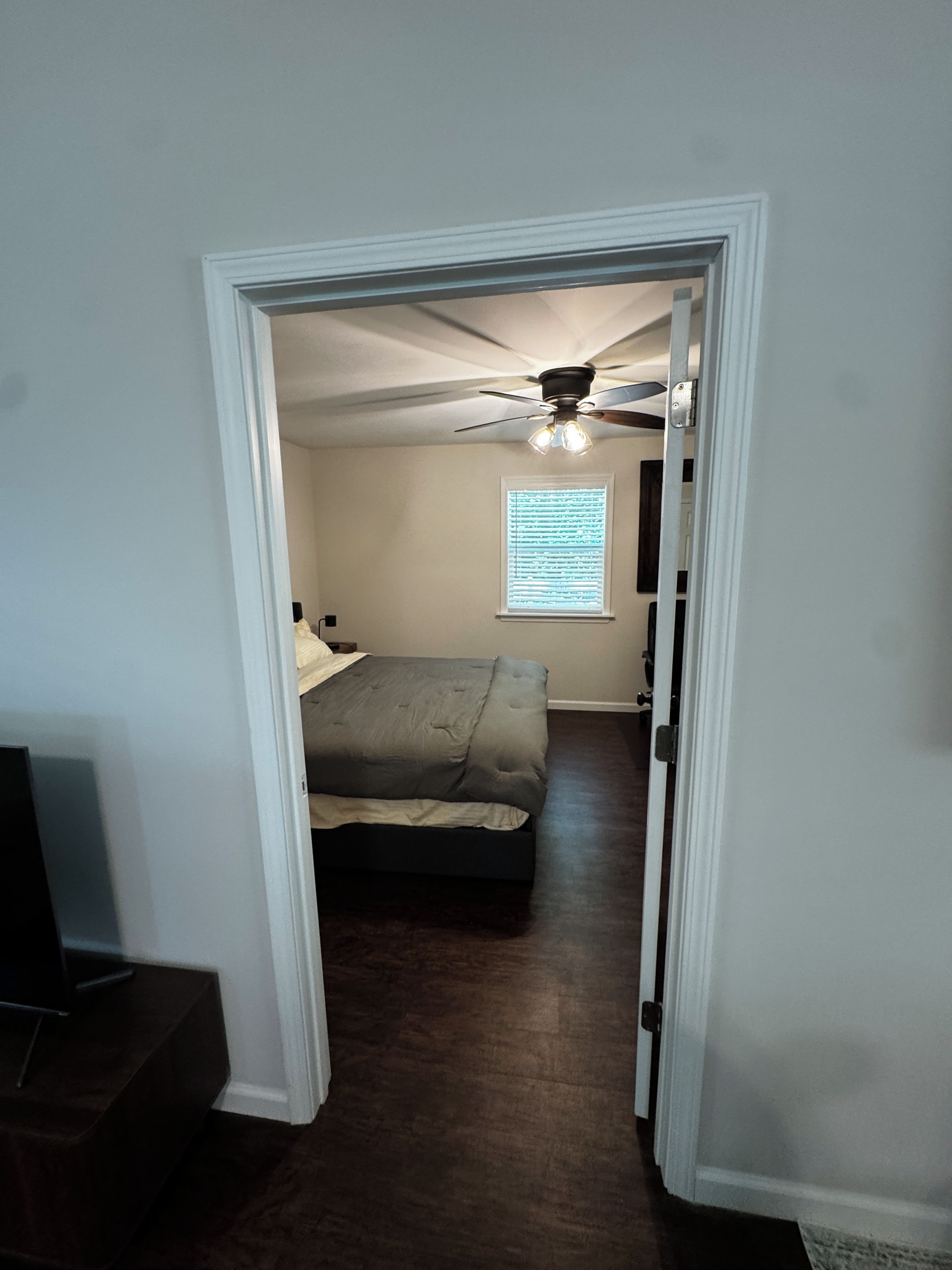 Une chambre à coucher comprenant un lit, un ventilateur de plafond, une fenêtre avec des stores et une porte donnant sur une autre pièce.