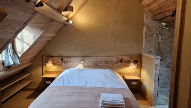 Ein Schlafzimmer mit schrägen Wänden, einem Holz-Kopfteil, zwei Nachttischlampen, einem Bett mit weißer Bettwäsche und einem sichtbaren Duschbereich.