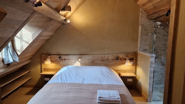 Ein Schlafzimmer mit schrägen Wänden, einem Holz-Kopfteil, zwei Nachttischlampen, einem Bett mit weißer Bettwäsche und einem sichtbaren Duschbereich.