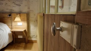 Una puerta de madera con un picaporte metálico, un pequeño escritorio de madera con una lámpara y una cama con cubrecama blanco.