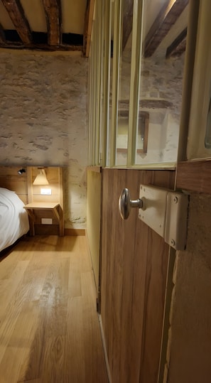 Eine Holztür mit einem Metallbeschlag, ein kleiner Holztisch mit einer Lampe sowie ein Bett mit weißer Bettdecke.