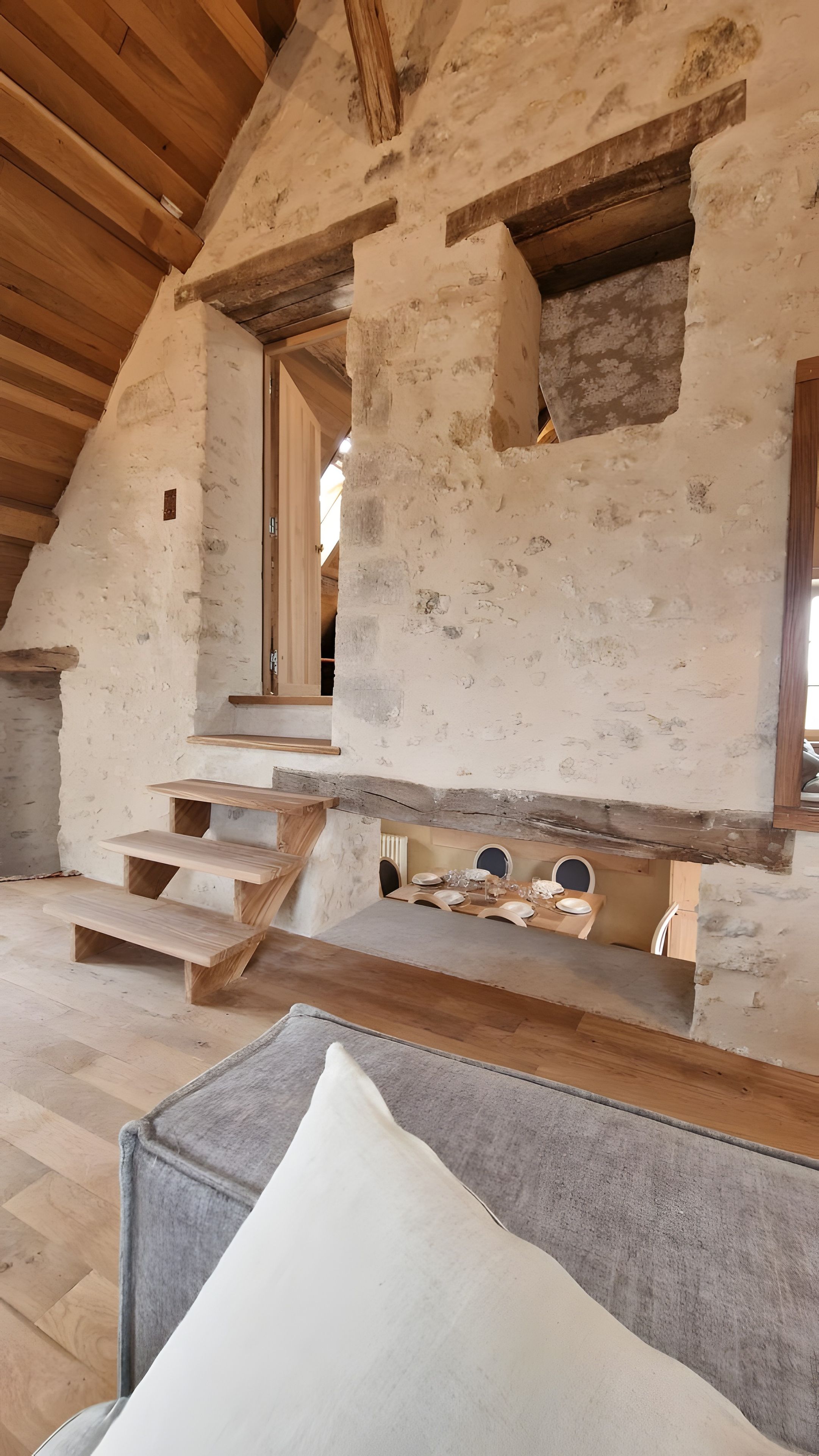 Un intérieur rustique avec un escalier en bois, un coin repas avec une table dressée pour quatre personnes et un espace salon confortable agrémenté d’un coussin.