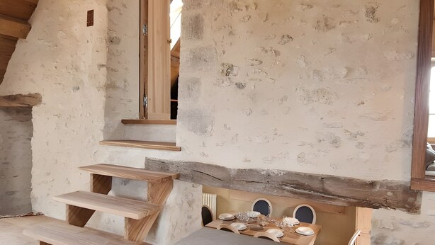 Un interior rústico con una escalera de madera, un comedor con una mesa dispuesta para cuatro personas y una zona de estar acogedora con cojines.