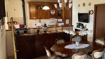 A dining area with a wooden table and chairs, a chandelier, and a kitchen in the background.