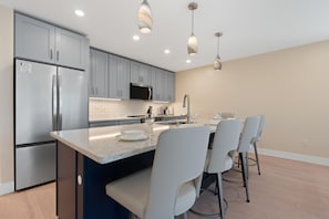 A modern kitchen with grey cabinets, a stainless steel refrigerator, a microwave, and a central island with bar stools. - Salty Seahorse (Fort Myers Beach)