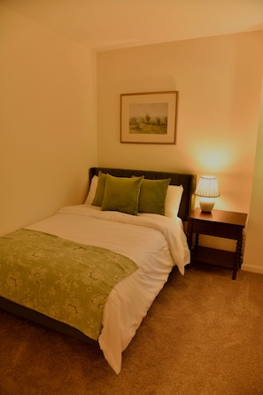 Une chambre à coucher avec un lit, des oreillers, une table de chevet avec une lampe, un tableau encadré au mur et un sol recouvert d’une moquette.