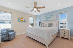 A bedroom with a bed, a chair, a nightstand, a ceiling fan, and artwork on the walls. - Salty Seahorse Unit 100 (Fort Myers Beach)