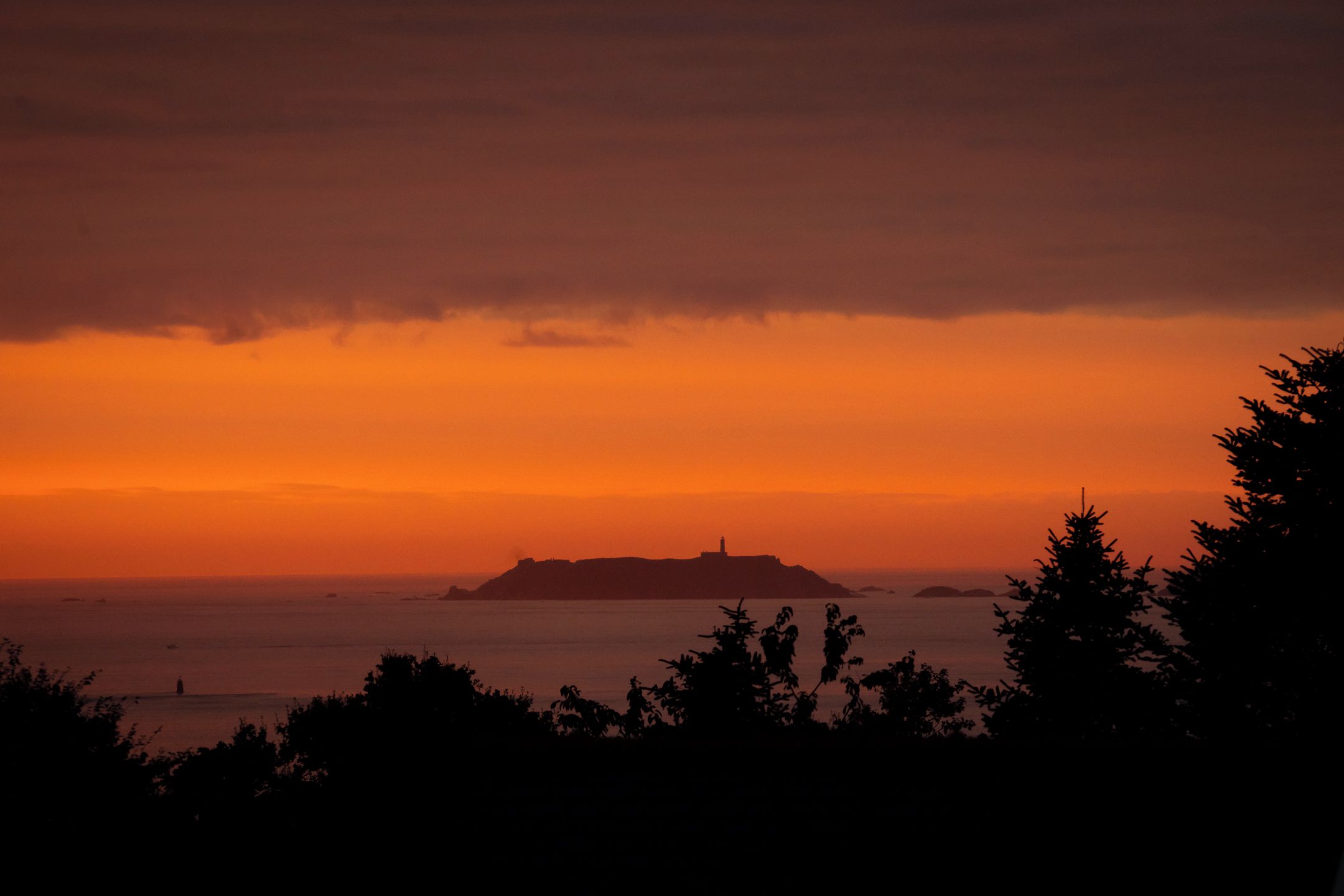 Paesaggio costiero con una collina che si distingue e la sagoma di un faro su uno sfondo di cielo al tramonto.