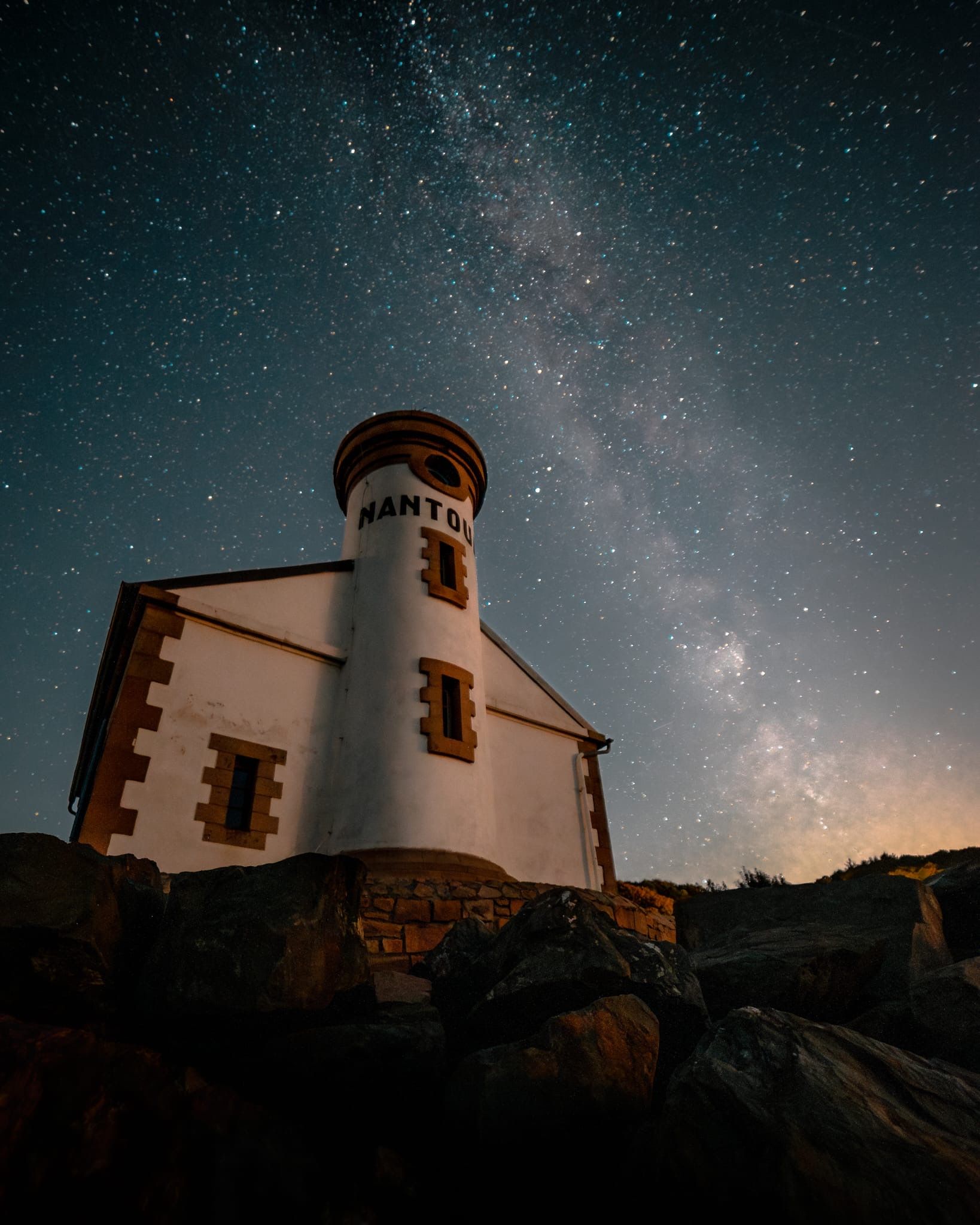Ein Leuchtturm mit dem Namen 'NANTOU' auf dem Turm, umgeben von Felsen unter einem sternenübersäten Nachthimmel.