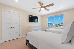 A bedroom with a bed, a dresser, a ceiling fan, a TV, and a window with a view of buildings. - Salty Seahorse Unit 200 (Fort Myers Beach)