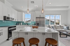 A modern kitchen with white cabinets, a central island with stools, stainless steel appliances, and a view of the outside through large windows. - Calico Jack's Lookout (Fort Myers Beach)