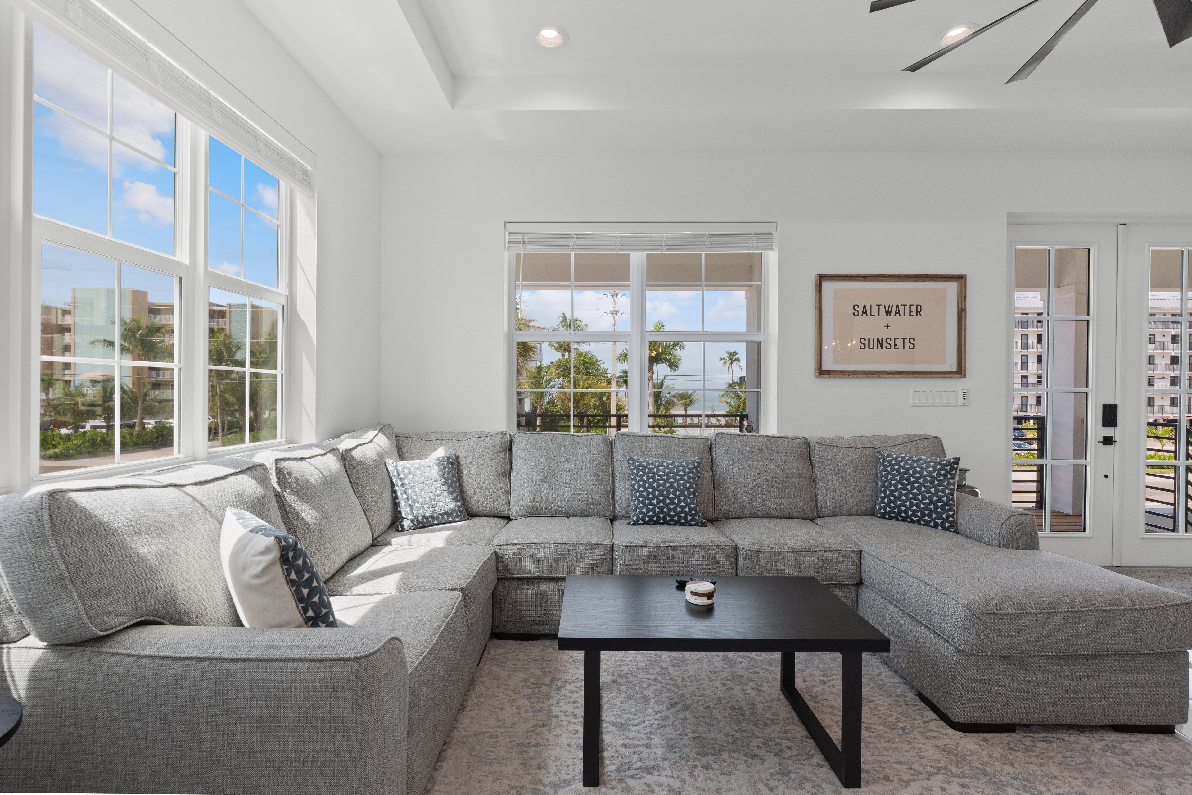 A modern living room with a sectional sofa, a coffee table, and large windows offering a view of palm trees and buildings.