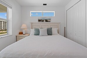 A bedroom with a large bed, a nightstand with a lamp, a window with a view of buildings, and a painting above the bed. - Calico Jack's Lookout (Fort Myers Beach)