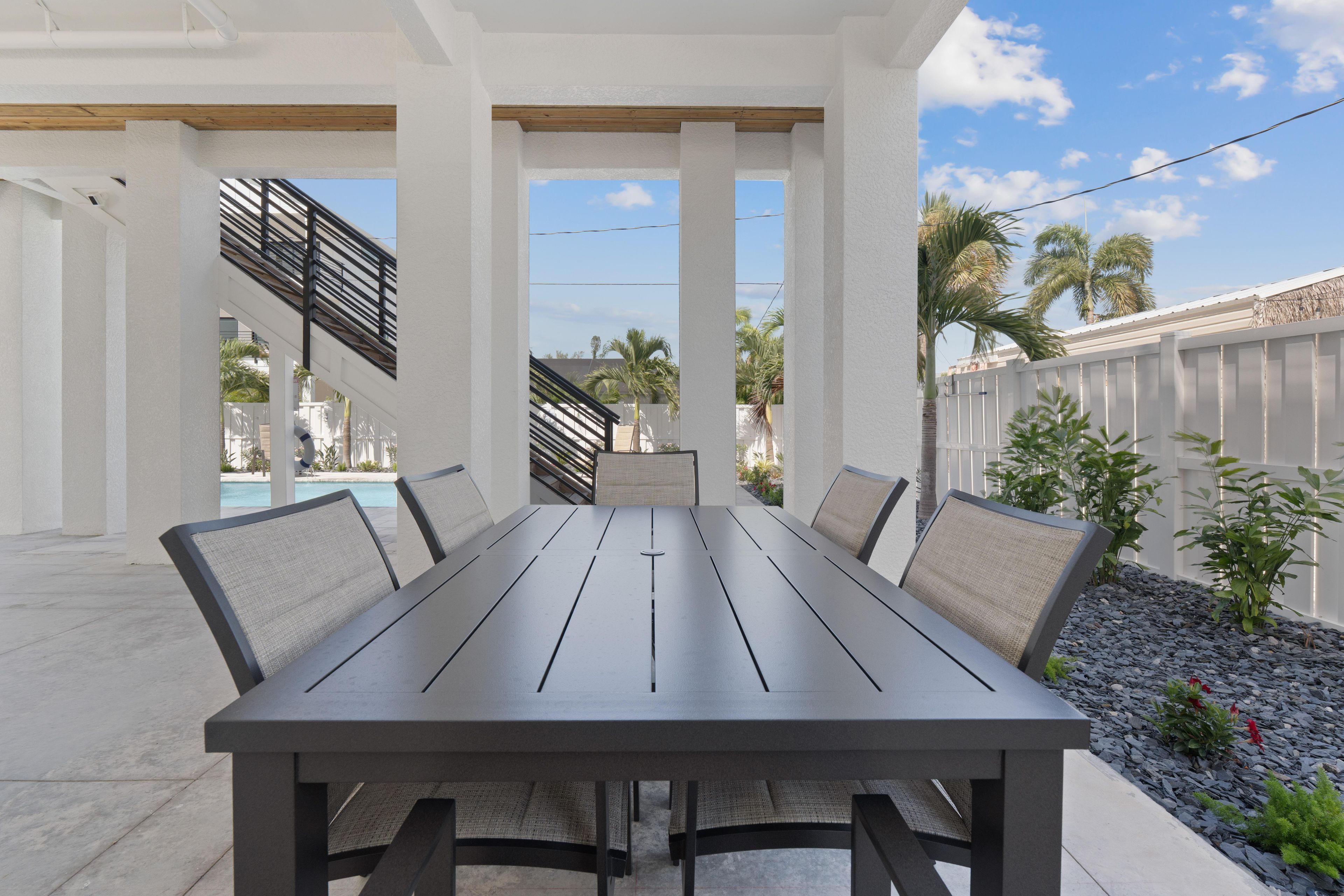 Een overdekt terras met een rechthoekige tafel en stoelen, een zwembad op de achtergrond en uitzicht op palmbomen en een hek.
