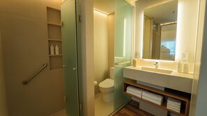 Un baño con cabina de ducha de vidrio, inodoro, lavabo con espejo y repisas con artículos de aseo.