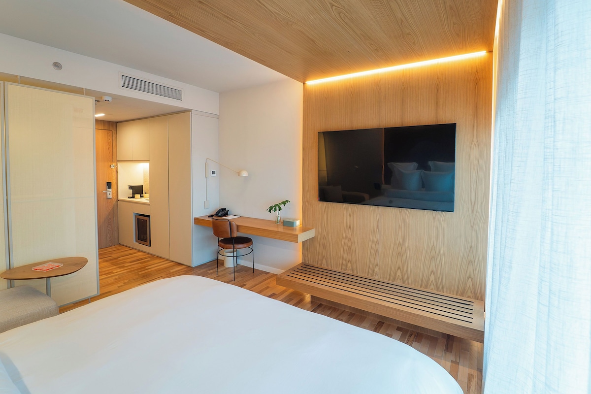 Habitación de hotel con una cama, un escritorio con silla, un televisor montado en la pared y una mesita con una planta.